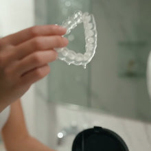 Gouttière dentaire transparente tenue à la main devant le nettoyeur Sonalys ouvert, dans une salle de bain
